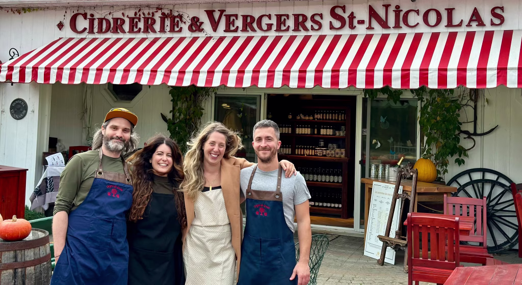Quatre membres souriants du personnel devant la Cidrerie & Vergers St-Nicolas avec une enseigne rouge et blanche, sous un auvent rayé rouge et blanc